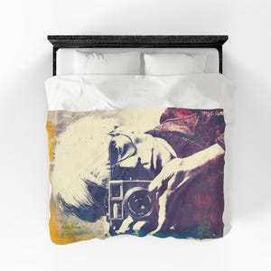 May include: A white duvet cover featuring a large, stylized graphic print of a person holding a vintage camera to their face. The artwork uses a distressed, multi-colored palette of yellow, blue, and deep red tones against a light background.