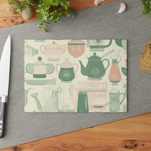 Pode incluir: Uma tábua de corte retangular de vidro com um padrão repetitivo de utensílios de cozinha, incluindo bules, açucareiros e jarros em tons de verde sálvia, rosa suave e creme, exibindo vários recipientes de cerâmica com tampas e pegas.
