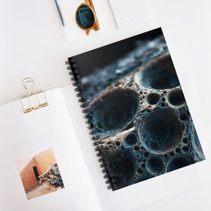 May include: Spiral-bound notebook with a unique, abstract design. The cover features a close-up view of a textured, porous surface in shades of blue, teal, and beige. The notebook is open, revealing blank white pages and a gold paper clip.