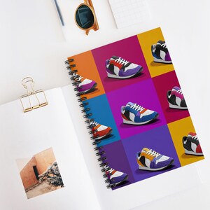 May include: Spiral-bound notebook featuring a colorful pop art design of sneakers. The sneakers are depicted in various colors, including red, purple, blue, and yellow, set against a vibrant background of squares. The notebook is open, revealing a blank page.
