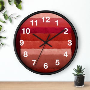 May include: A round wall clock with a black frame. The clock face features horizontal bands of red hues, from light to dark. White numerals mark the hours. Black hands indicate the time.