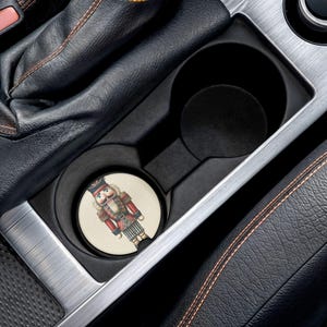 Puede incluir: Un posavasos para coche con diseño de cascanueces. El posavasos redondo tiene un fondo crema y una ilustración detallada de un cascanueces con uniforme rojo y negro. El posavasos está colocado en el portavasos de un coche.