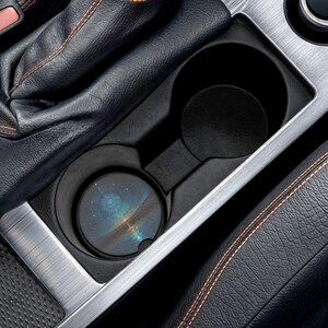 May include: A circular car cup holder coaster featuring a dark blue and orange celestial nebula print with stars and a glowing horizon line, placed inside a black plastic vehicle center console cup holder next to a black leather gear shift boot.