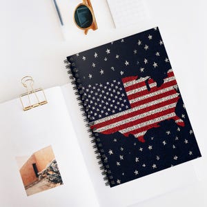 May include: Spiral-bound notebook with a design of the American flag and a map of the United States. The cover has a navy blue background with white stars. The flag and map are in red and white. The notebook is open on a white surface.
