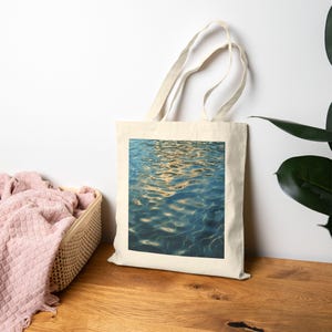 May include: A cream-colored canvas tote bag featuring a centered square graphic print of rippling blue water with golden light reflections. The bag has two long shoulder straps and a rectangular body made of a woven fabric material.