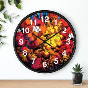 Puede incluir: Un reloj de pared redondo con marco negro. La esfera presenta un diseño abstracto y colorido que se asemeja a una explosión en tonos rojos, naranjas, amarillos y azules. Los números blancos marcan las horas.