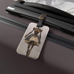 Puede incluir: Una etiqueta de equipaje rectangular con una ilustración gráfica de una figura humanoide mecánica con articulaciones en tono bronce y una falda en capas de color crema. La etiqueta está sujeta a un asa de maleta gris oscuro con una correa.