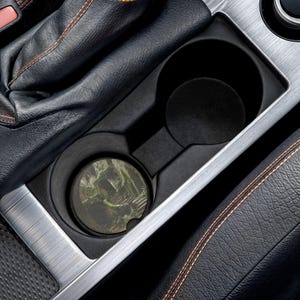 Puede incluir: Un posavasos circular para portavasos de coche con una imagen impresa de una escalera de piedra rodeada de follaje verde musgo y sombras oscuras, colocado dentro de un portavasos de consola central de plástico negro de un vehículo.