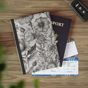 May include: A floral passport holder with a black border, a dark blue passport, and two boarding passes are on a wooden surface. The passport holder features a black and white floral design. A car key is also present.