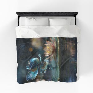 May include: A white duvet cover featuring a printed graphic of a translucent blue frog-like creature with crystalline textures positioned next to a pale pink water lily flower against a dark, moody, and blurred background with soft golden light.