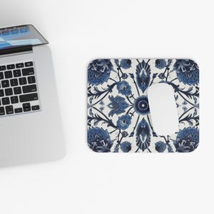May include: A rectangular mousepad with a white background and a blue floral pattern. A white computer mouse is on the mousepad. A laptop is in the upper left corner.