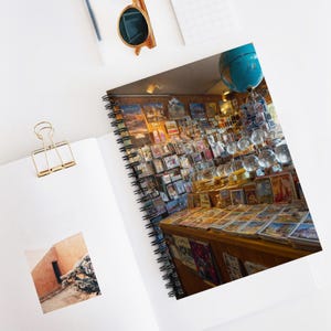 May include: A spiral-bound notebook with a photograph of a shop filled with postcards, globes, and glass spheres. The shop's interior is lit with warm lighting, and the walls are covered with various postcards. A pair of sunglasses and a gold paper clip are on the white surface.