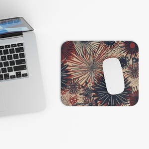 Pode incluir: Um mousepad retangular com um fundo creme e um padrão repetitivo de desenhos de explosões de estrelas vermelhas e azul marinho. Um mouse de computador branco repousa sobre a almofada, ao lado de um laptop.