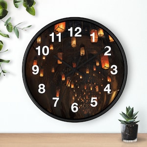 May include: Round black wall clock with a dark background featuring hanging lanterns. The clock face has white numerals and black hands. The clock is on a wooden surface with a plant in a pot.