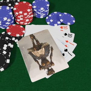 Puede incluir: Un mazo de cartas y pilas de fichas de póquer rojas, azules y negras. Una carta muestra una ilustración invertida de una figura mecánica con extremidades metálicas y una falda texturizada. Las otras cartas visibles muestran un diez y una sota.