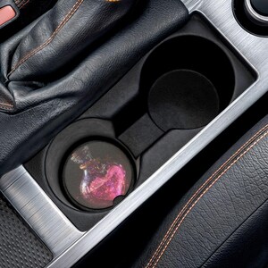 以下が含まれることがあります： 黒い内装とシルバーのトリムが付いた車のカップホルダー。ピンクと紫のデザインの丸いコースターがカップホルダーに入っています。コースターは、ハート型の中心部を備えたボトルのデザインになっています。