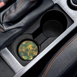 Puede incluir: Un posavasos circular para portavasos de coche con una vista aérea de un paisaje de mosaico con varios tonos de campos verdes, amarillos y marrones. El posavasos está colocado dentro de una consola de portavasos de plástico negro.