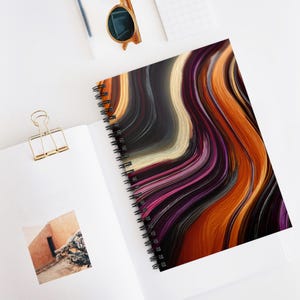 May include: Spiral-bound notebook with a colorful abstract design. The cover features flowing lines in shades of orange, purple, black, and cream. The notebook is open, revealing white pages, and rests on a white surface with a gold paper clip and sunglasses.