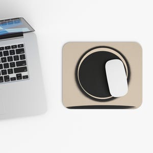 Könnte beinhalten: Ein beigefarbenes rechteckiges Mauspad mit einem schwarzen Kreisdesign und einer weißen Computermaus. Das Mauspad hat einen schwarzen Sprühfarbeffekt um den Kreis herum. Ein Laptop ist teilweise in der oberen linken Ecke sichtbar.