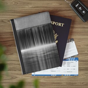 May include: A black and gray passport holder with a landscape design, a dark blue passport, and two airline tickets are on a wooden surface. The passport holder has a textured design. A car key is also present.