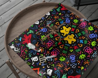May include: A wrapped gift box with a brown ribbon sits on black wrapping paper with a colorful, abstract pattern. The paper features shapes, symbols, and figures in red, yellow, green, blue, and orange. A roll of tape and scissors are also on the paper.