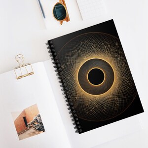 Può includere: Quaderno a spirale nero con un design geometrico dorato sulla copertina. Il design è una serie di cerchi e linee concentriche, che creano un effetto vortice. Il quaderno è aperto su una superficie bianca, con una graffetta dorata e occhiali da sole.