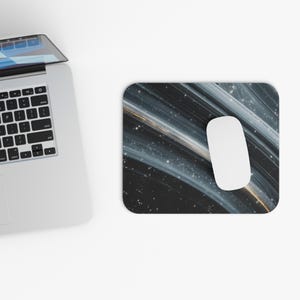May include: A white computer mouse sits on a rectangular mousepad. The mousepad has a dark background with flowing white and gold lines, and small white dots. A laptop is partially visible in the upper left corner.