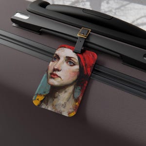 May include: A rectangular luggage tag with a painted portrait of a woman with blue eyes and a red headscarf. The tag is attached to a black suitcase with a gold-coloured buckle.