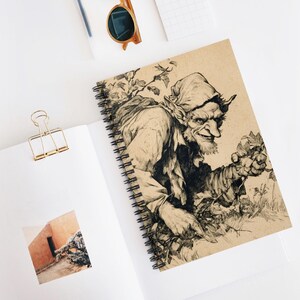 Puede incluir: Cuaderno de espiral con una ilustración detallada en blanco y negro de un personaje sonriente con orejas puntiagudas y un sombrero. La obra de arte está sobre un fondo beige, con una encuadernación en espiral negra. El cuaderno está abierto sobre una superficie blanca.