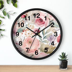 May include: A round wall clock with a black frame and a white face featuring a watercolor floral pattern of pink roses, green leaves, and muted purple accents. The clock has black hour, minute, and second hands with bold black numbers 1 through 12.