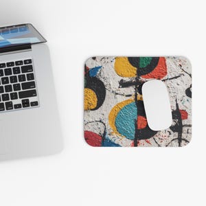 May include: A white computer mouse rests on a rectangular mousepad with an abstract design. The mousepad features a textured, off-white background with colorful shapes in yellow, blue, red, green, and black. A laptop is partially visible in the upper left corner.