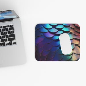 May include: A rectangular mousepad with a vibrant, iridescent scale pattern in shades of blue, purple, and green. A white computer mouse rests on the pad, next to a laptop.
