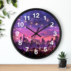May include: A round wall clock with a black frame featuring a printed image of a city skyline at sunset with purple and pink clouds. The clock face displays white numbers from 1 to 12 and black hour, minute, and second hands over a string light design.