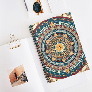 Peut inclure: Cahier à spirales avec une couverture ornée d'un mandala coloré. Le motif présente des éléments turquoise, rouges et dorés disposés en cercles concentriques. Le cahier est ouvert sur une surface blanche, avec une pince à papier dorée et des lunettes de soleil.