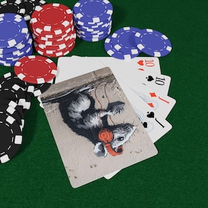 Puede incluir: Una imagen con temática de póquer con pilas de fichas rojas, azules y negras sobre una superficie de fieltro verde. Un mazo de cartas está extendido, con una carta que presenta una rata con auriculares. Las cartas muestran el número 10.