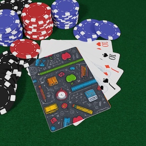 May include: Poker chips in black, red, and blue are stacked on a green surface, with a deck of cards fanned out. One card features a school-themed design with pencils, apples, and a ruler.