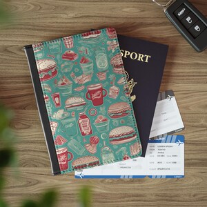 May include: A teal passport holder with a black border, featuring a repeating pattern of burgers, fries, and drinks in red and white. A navy blue passport, plane tickets, and car keys are also visible on a wooden surface.