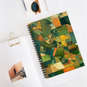 Puede incluir: Cuaderno de espiral con una vista aérea colorida de campos agrícolas en tonos verdes, amarillos, naranjas y marrones. El cuaderno está sobre una superficie blanca, con un clip dorado y gafas de sol.