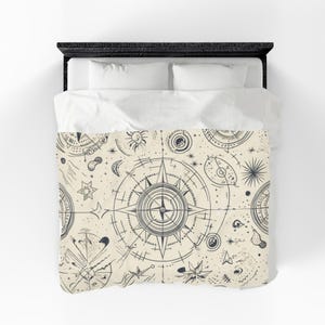 May include: Cream-coloured duvet cover with a celestial design. The pattern features compasses, stars, moons, and other astronomical elements in dark blue. The bedding is on a black bed frame with white pillows.