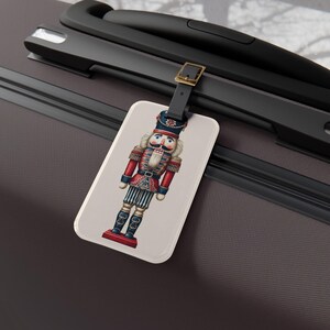 May include: A luggage tag featuring a detailed illustration of a traditional nutcracker soldier. The tag has a cream background and a black leather strap with a gold buckle. The nutcracker is in red, blue, and white.