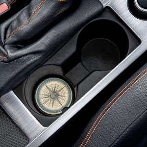 May include: A car cup holder with a compass design coaster. The coaster has a vintage map design with a compass rose in the center. The cup holder is black and silver, and the car interior is black leather.