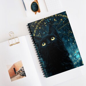 May include: A spiral-bound notebook with a black cat illustration on a dark blue background with gold details. The notebook is open on a white surface, with a gold paper clip and sunglasses visible. The cat has yellow eyes.