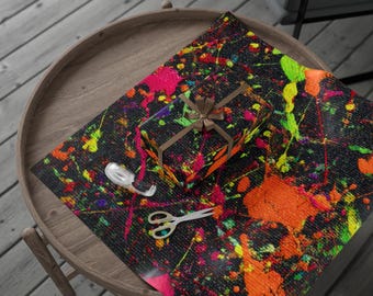 May include: A wrapped gift box with a brown ribbon sits on black wrapping paper covered in colorful paint splatters. A roll of tape and scissors are also on the wrapping paper. The items are on a round wooden table.