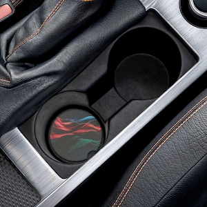 May include: A black car cup holder with a coaster featuring a colorful abstract design of red, blue, and green wavy lines. The cup holder is set in a silver and black interior.