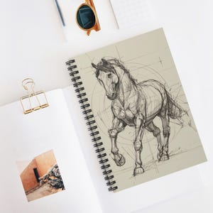 May include: Spiral-bound notebook featuring a detailed pencil sketch of a running horse. The drawing is on a beige background with faint geometric construction lines. The notebook has a black spiral binding.