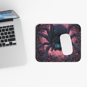 May include: A computer mouse and a mousepad featuring a detailed illustration of a large, dark blue spider with a glowing blue center. The background is a mix of pink and purple hues.