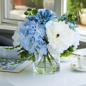 May include: A bouquet of artificial flowers in a clear glass vase. The arrangement includes white and pale blue hydrangeas, blue dahlias, and green leaves. The vase is on a white surface, next to a magazine and a teacup.