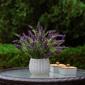 May include: A white ceramic vase with a textured vertical stripe design holds a bouquet of purple lavender. Two small white square containers with wooden lids sit nearby on a glass-topped table. The background is a blurred view of green foliage.