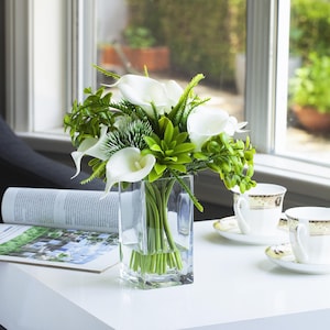 Op de afbeelding: Een heldere, vierkante glazen vaas met een boeket witte calla lelies en groen blad. De opstelling staat op een witte tafel, met kopjes en een tijdschrift. Natuurlijk licht komt door een raam.