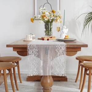 May include: A wooden dining table is set with a white lace table runner. The table is decorated with a vase of flowers, a cup and saucer, a plate, and a candle holder. Two wooden chairs with woven seats are in the foreground.